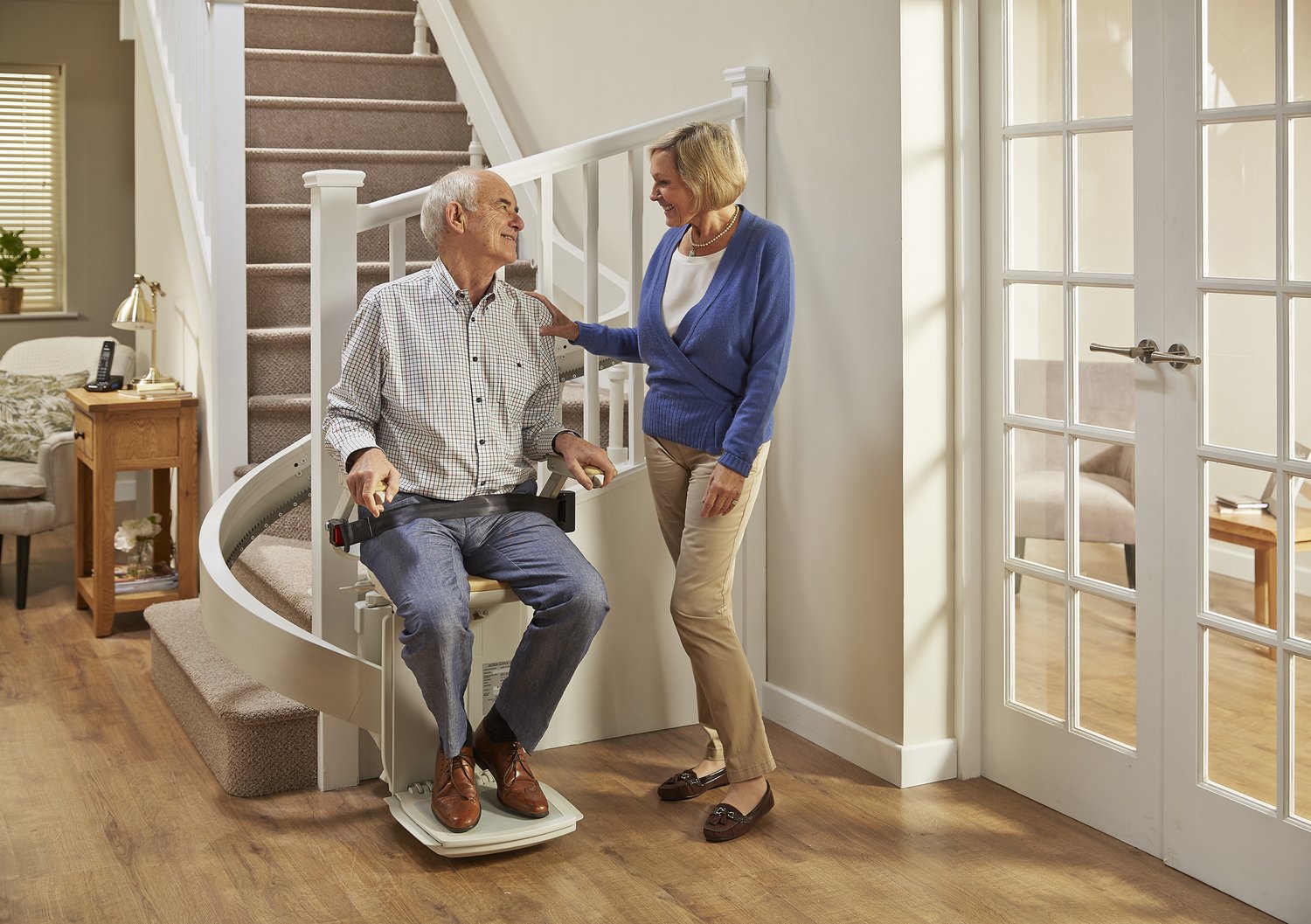 A wife and her husband at the stairs with the elderly man sitting on a stair lift